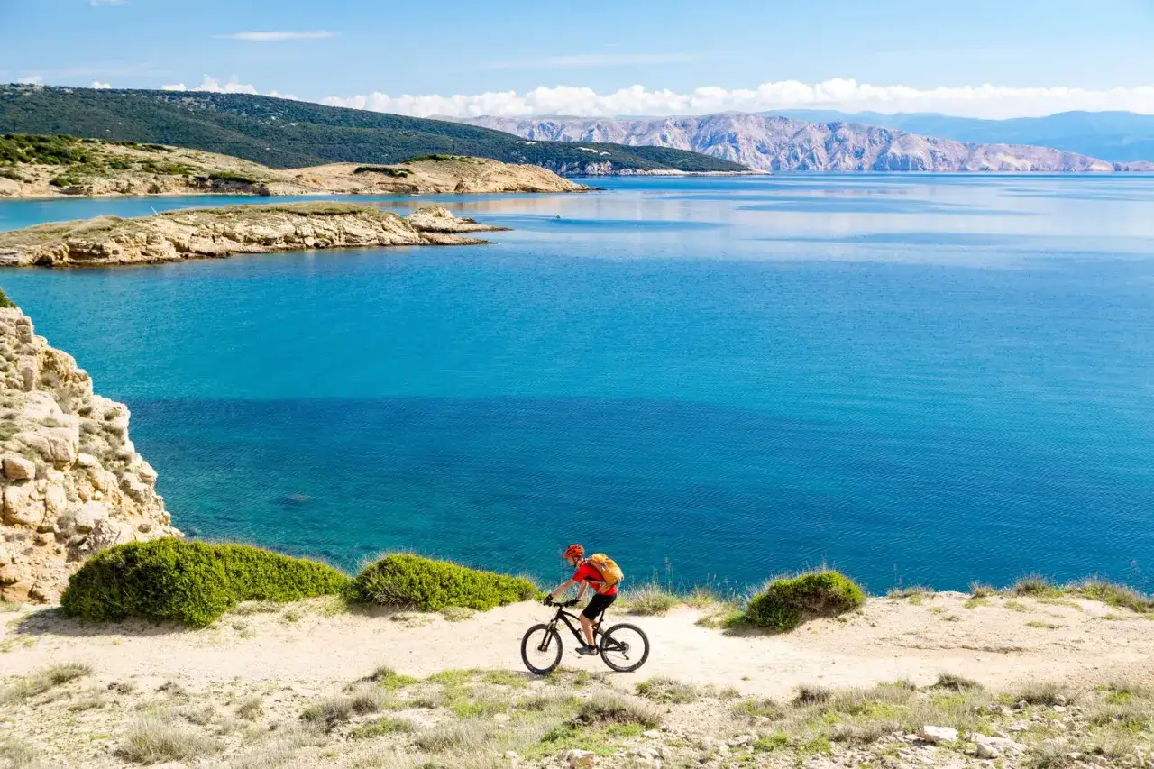 Ciclista disfruta de un recorrido en Croacia en bicicleta, con el mar turquesa y montañas de fondo.