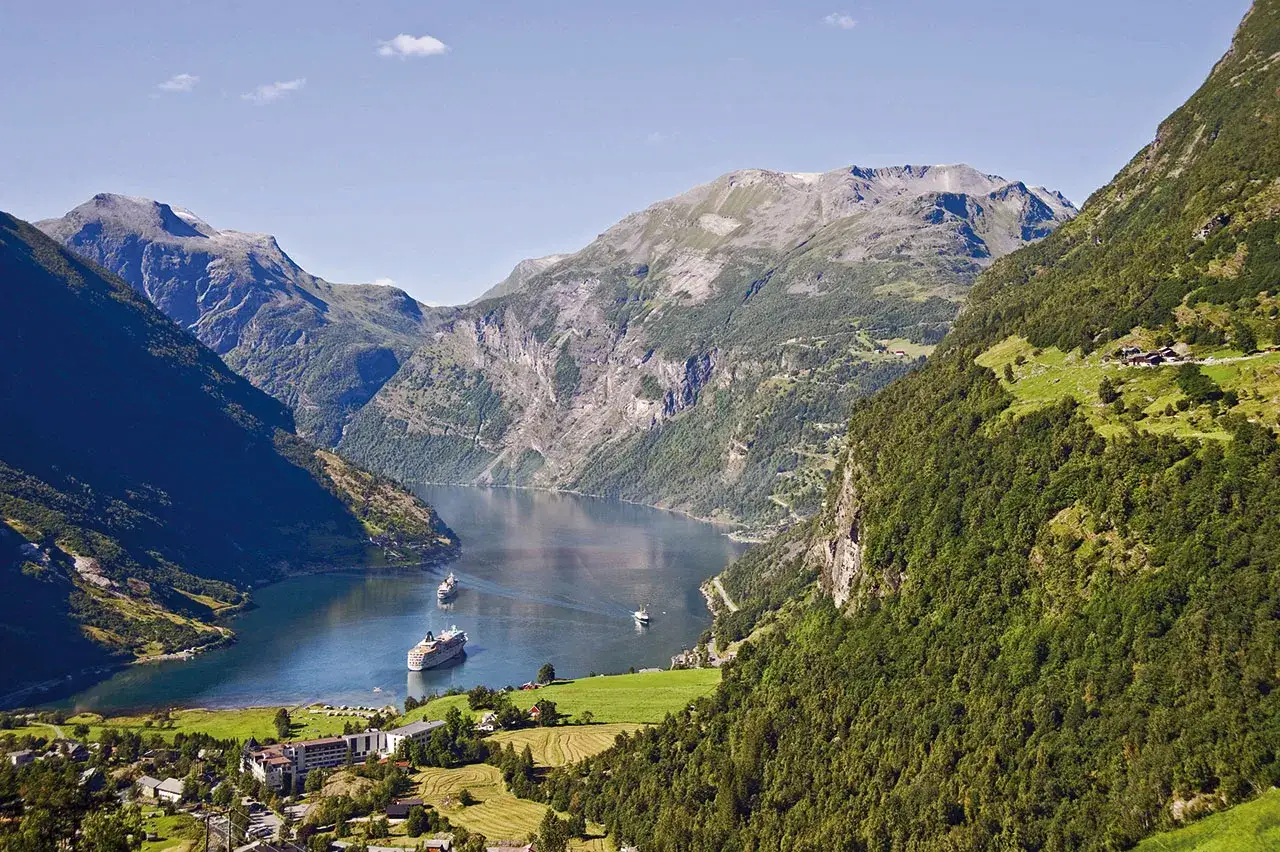 Piękna pogoda w Geiranger. Statki wycieczkowe płyną po spokojnym fiordzie, otoczonym zielonymi górami.