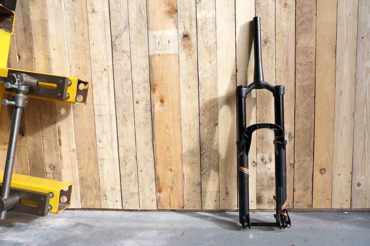 A black MRP Ribbon coil suspension fork stands against a wooden wall. The fork features orange accents and the