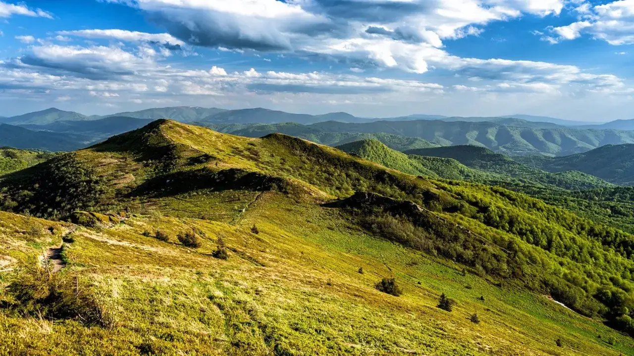 Zielone, pofałdowane wzgórza Bieszczadów pod błękitnym niebem z chmurami. Szlak wije się po zboczu, zapraszając do wędrówki. Gdzie w Bieszczady? Tu!
