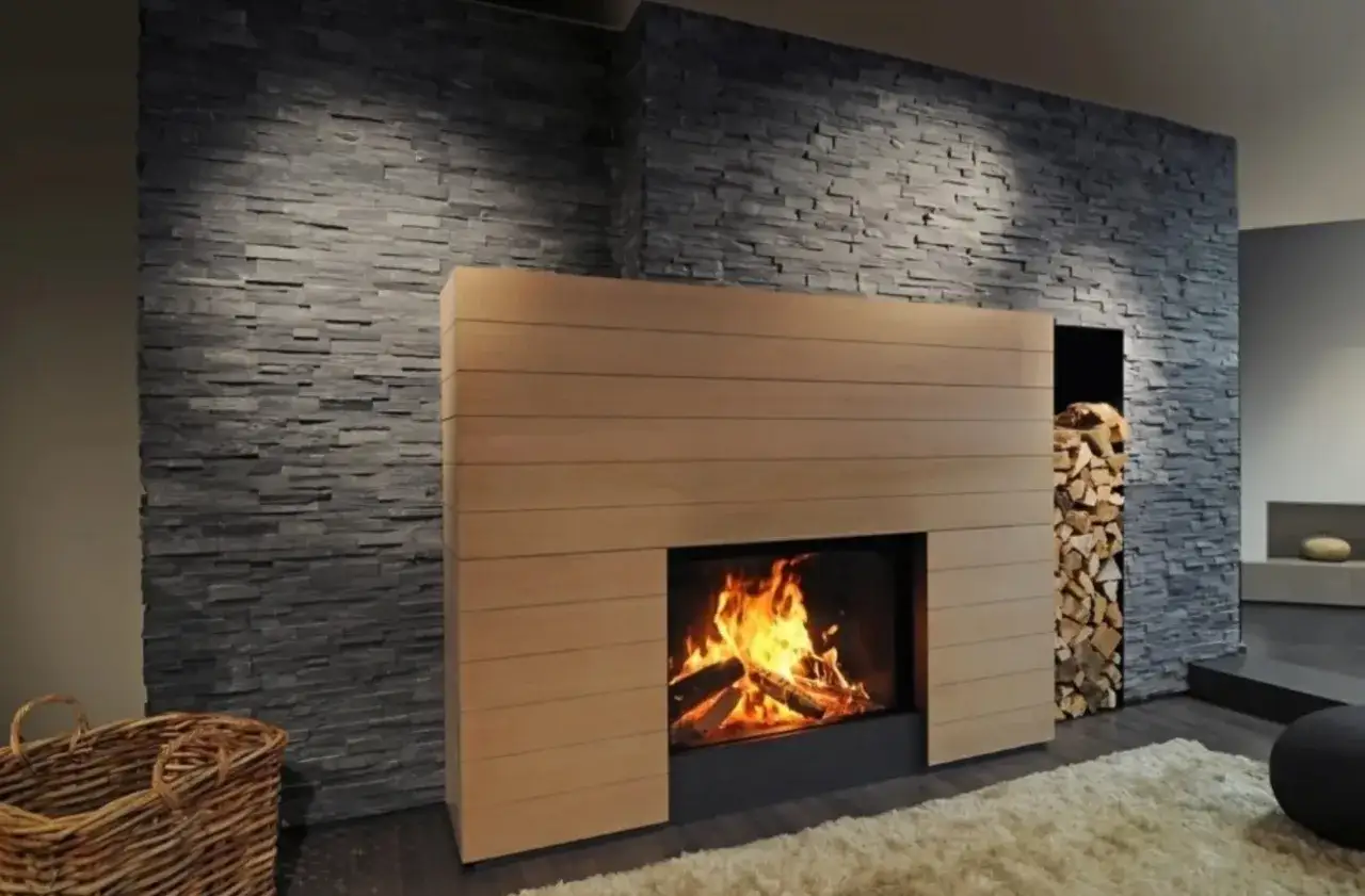 Camino fai da te in muratura con rivestimento in legno e pietra. Legna accatastata a lato, un cesto di vimini e un tappeto soffice completano l'ambiente.
