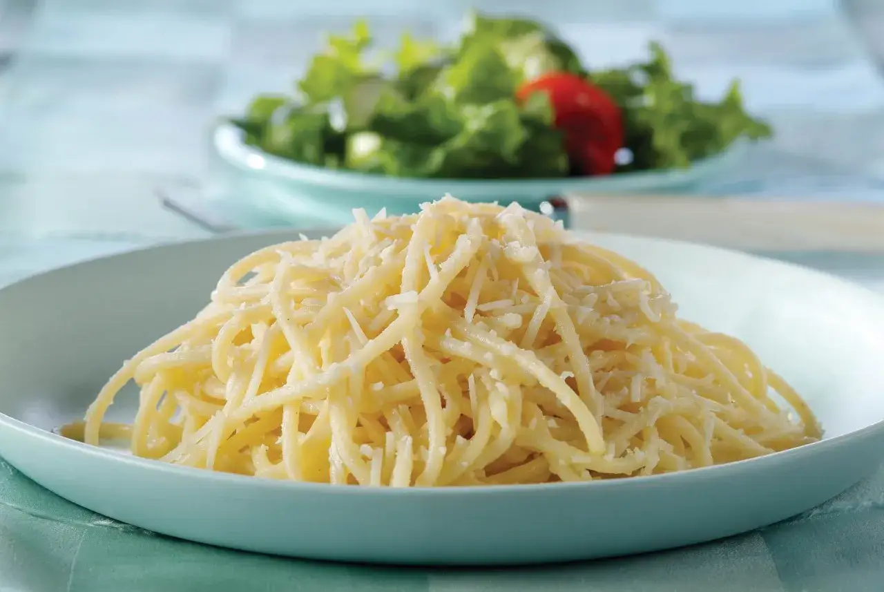Porcja spaghetti posypana obficie parmezanem startym, w tle sałatka z pomidorem.
