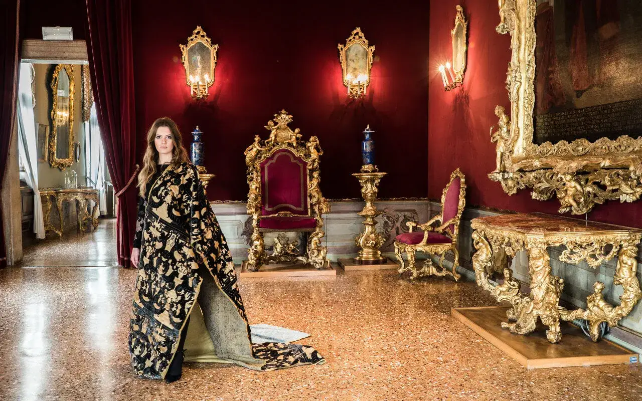 Donna con sontuoso abito in tessitura Bevilacqua Venezia, in una sala dorata con troni e specchi.