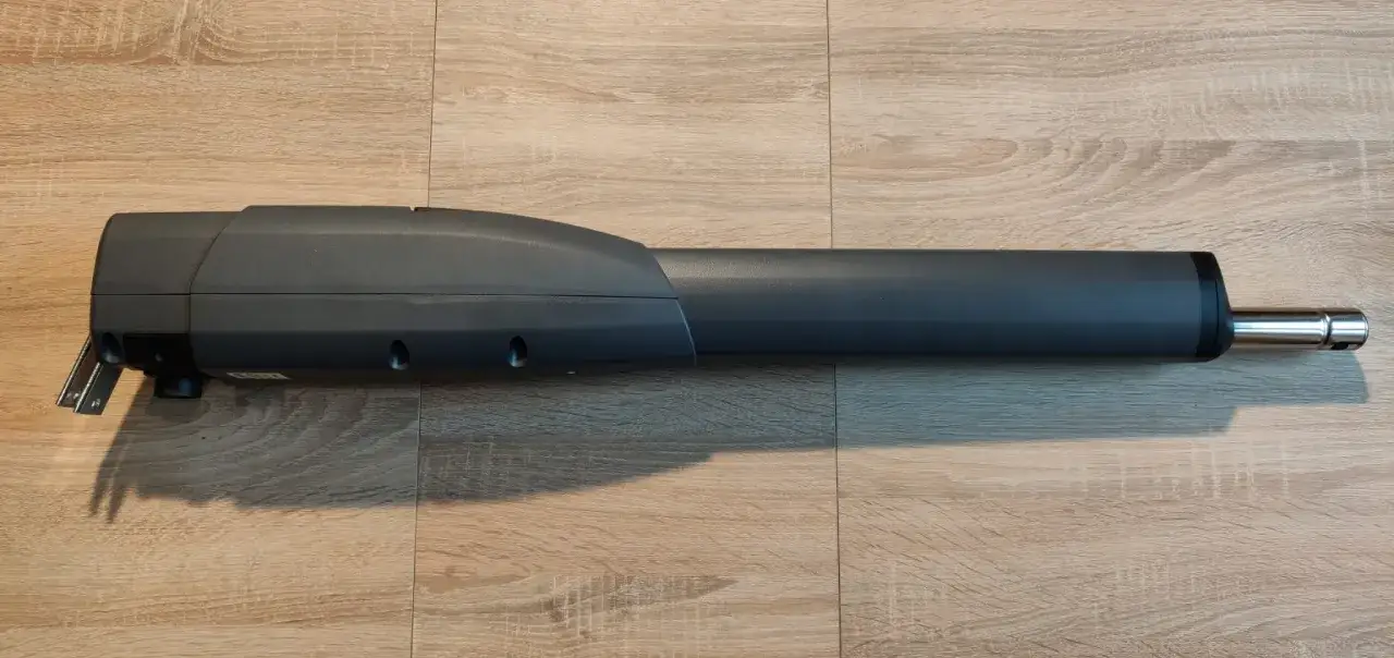 Siłownik do bramy przesuwnej, model ATS 24V, z metalowym wysięgnikiem, leży na drewnianej podłodze.