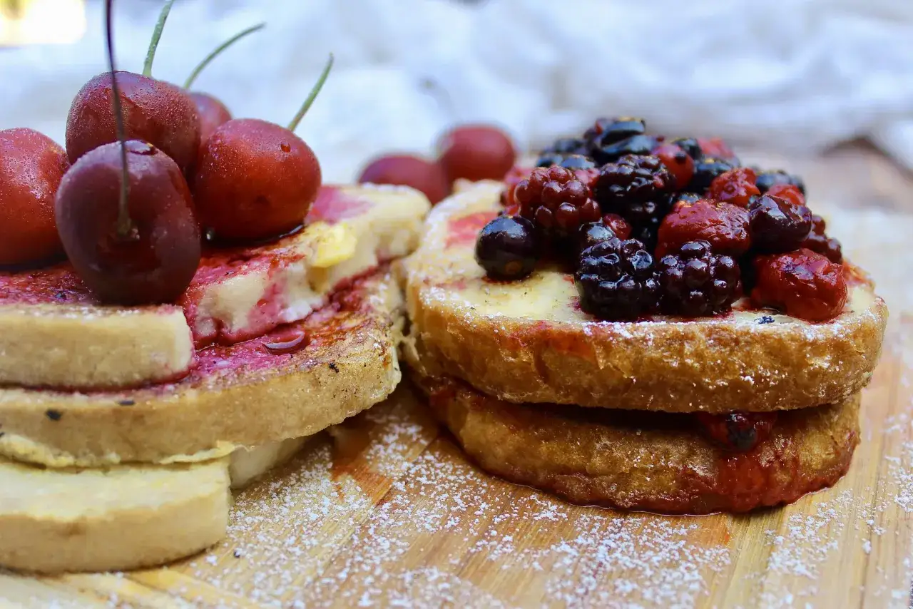 Zwei goldbraune Scheiben Pain Perdu, &uuml;ppig belegt mit frischen Kirschen und Beeren, best&auml;ubt mit Puderzucker.