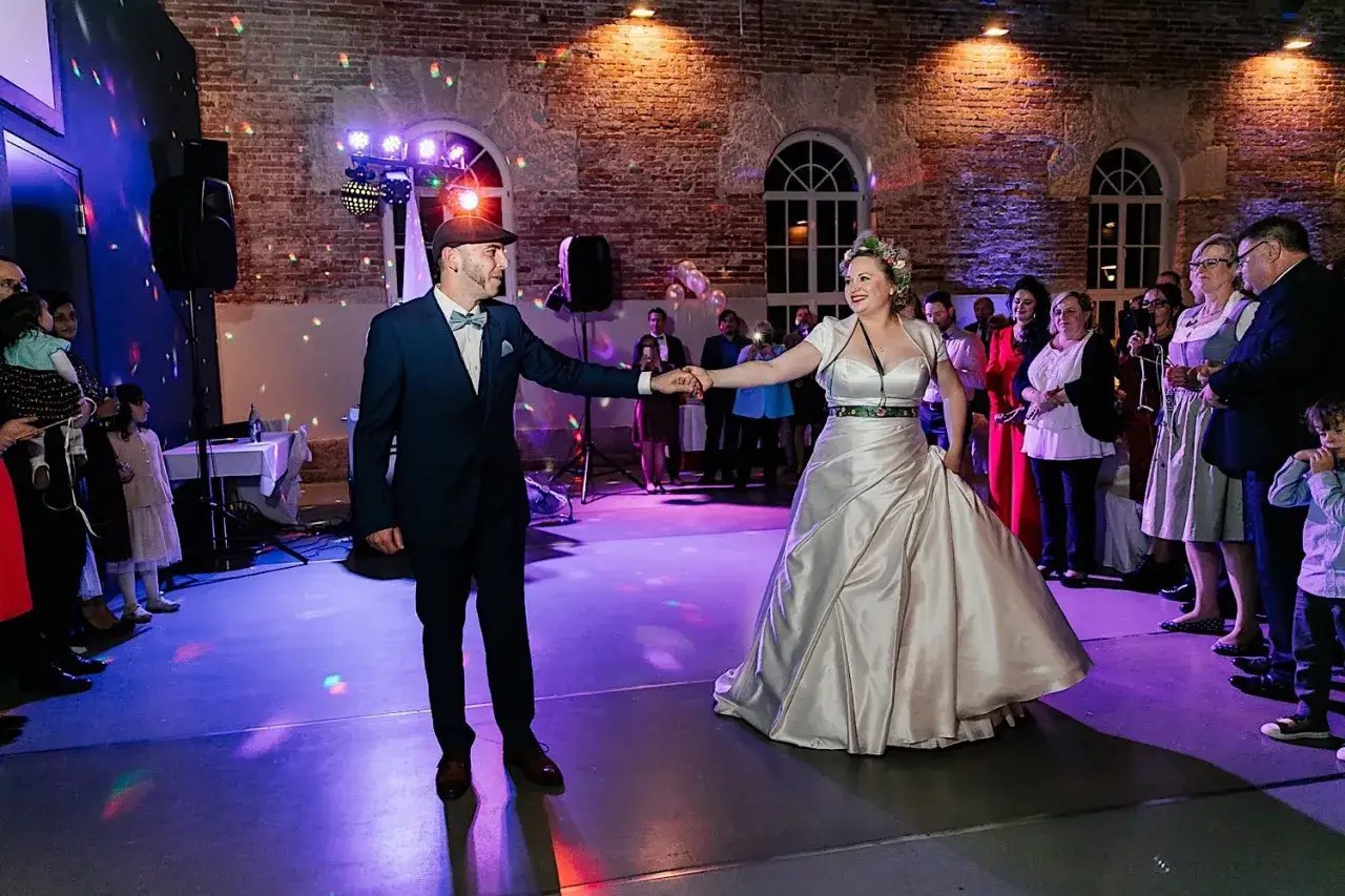 Brautpaar übt den Hochzeitstanz. Sie halten Händchen und lächeln sich an, während Gäste zusehen. Bunte Lichter tanzen auf dem Boden.