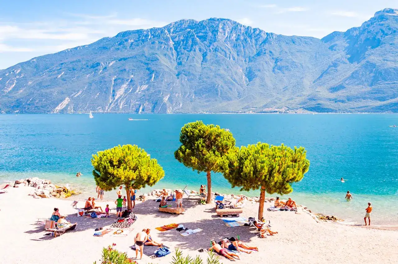 Sonnige Tage am Gardasee: Menschen entspannen am Strand, einige genießen das Gardasee schwimmen im klaren Wasser vor majestätischer Bergkulisse.