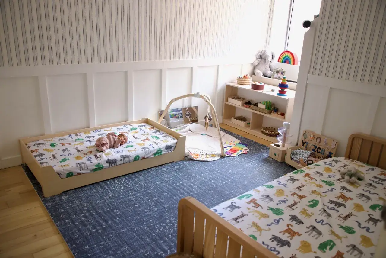 A baby sleeps on a floor bed with animal print sheets, showing how to make a floor bed for baby. A play mat and shelves with toys are nearby.