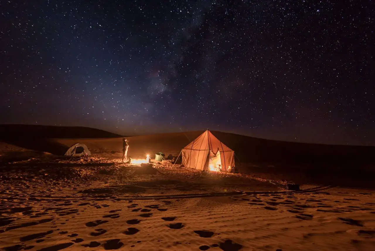 Une nuit dans le désert marocain, sous un ciel étoilé. Un campement avec une tente illuminée et un feu de camp réchauffe le sable.