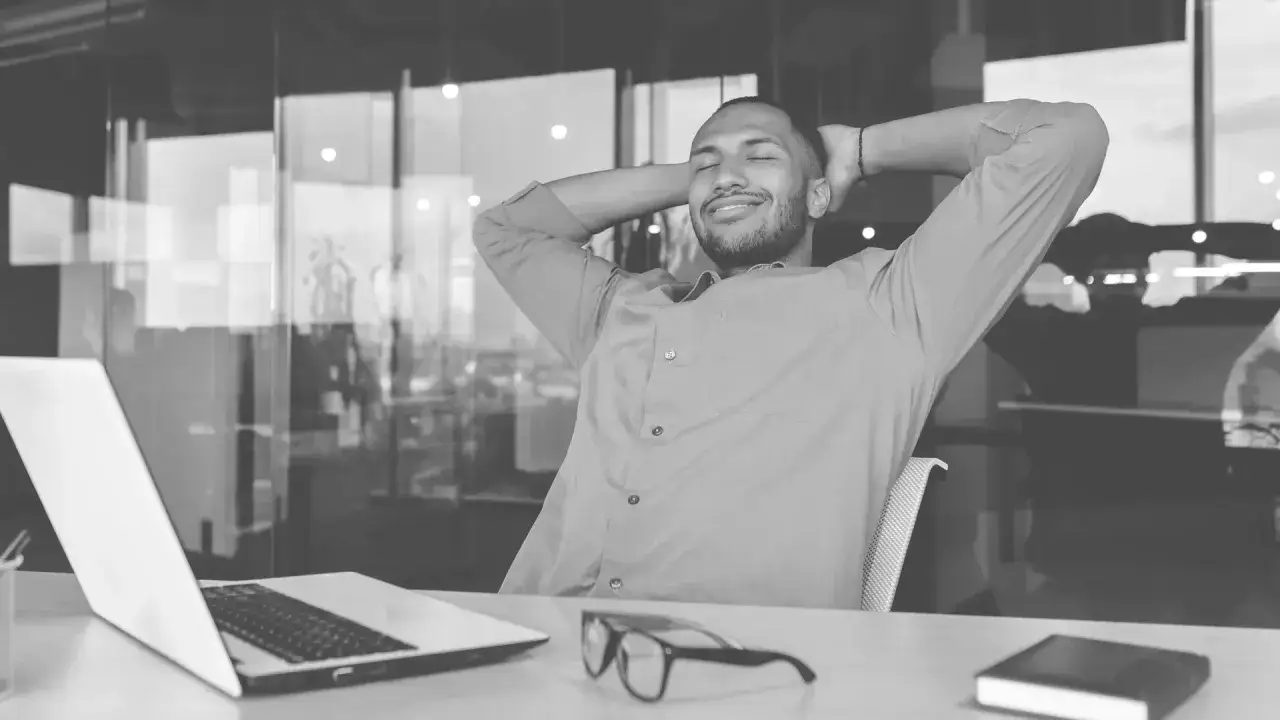 A man relaxes at his desk, hands behind his head, smiling. This is how to not be overwhelmed.