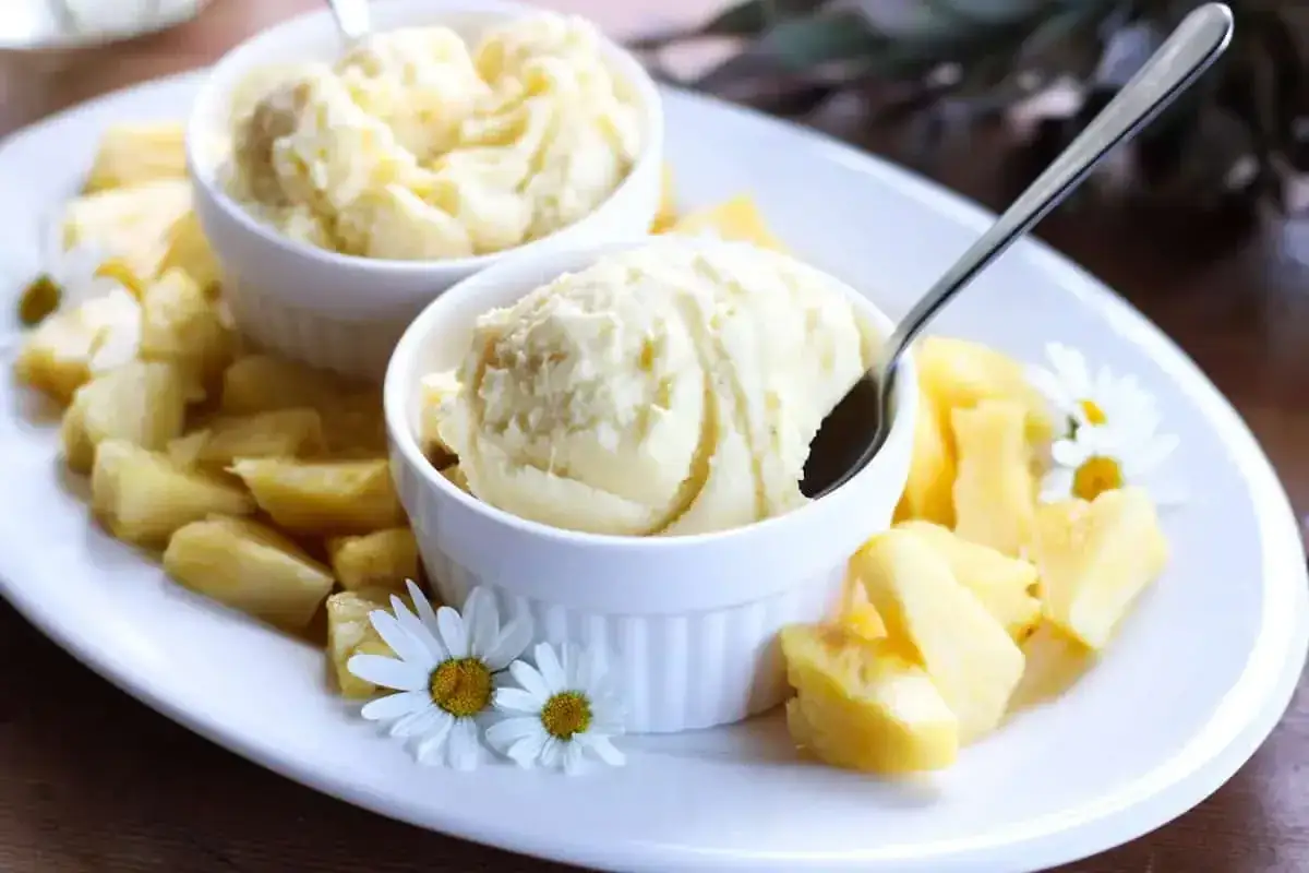 Zwei Schalen cremiges Ananas Eis, garniert mit frischen Ananasstücken und Gänseblümchen, auf einem weißen Teller.