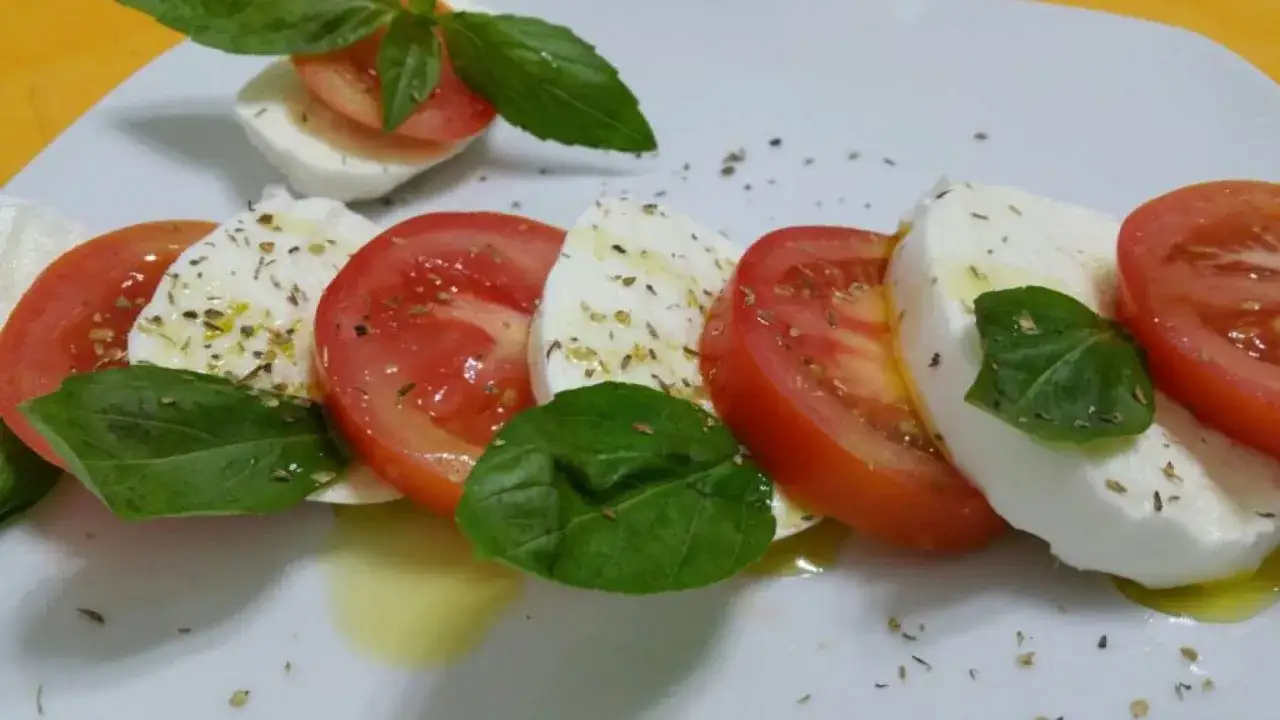 Insalata caprese: fette di pomodoro rosso e mozzarella bianca alternate, condite con olio, basilico fresco e origano.