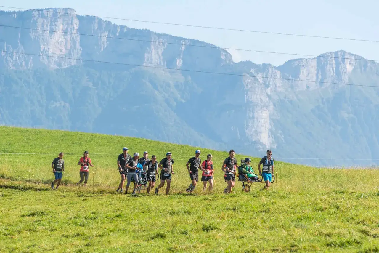 Une &eacute;quipe court solidaire lors d'une joelette course, traversant une prairie verdoyante avec des montagnes majestueuses en arri&egrave;re-plan.