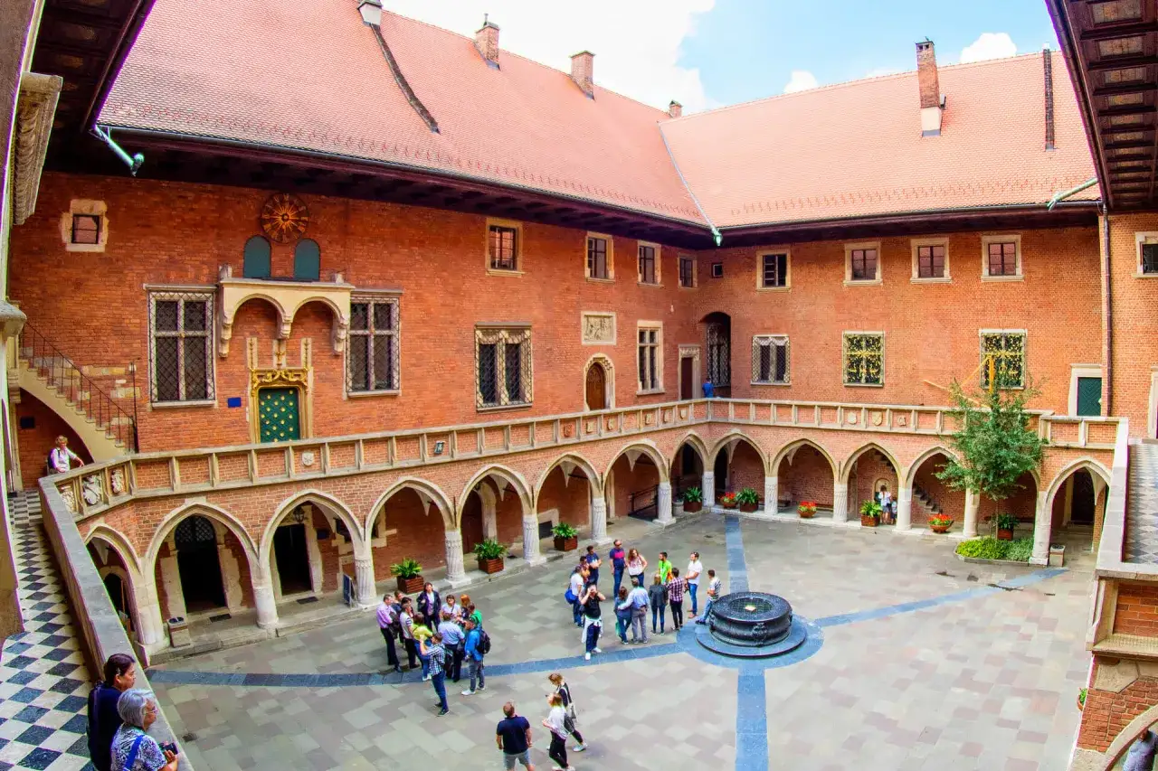 Dziedziniec Collegium Maius, z ceglanymi arkadami, schodami i grupami turyst&oacute;w zwiedzających historyczny budynek Uniwersytetu Jagiellońskiego.