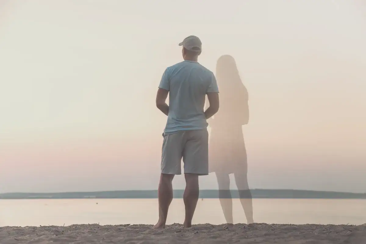 Uomo in piedi sulla spiaggia, con la figura evanescente di una donna al suo fianco. Un'immagine che evoca il sentirsi soli in coppia.
