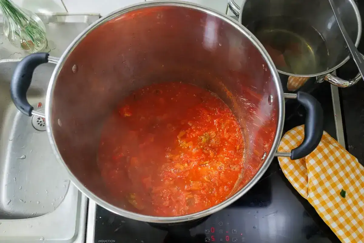 Stor gryta med bubblande tomats&aring;s, perfekt f&ouml;r ett hemlagat tomats&aring;s recept. En gulrutig grytlapp vilar p&aring; spisen.