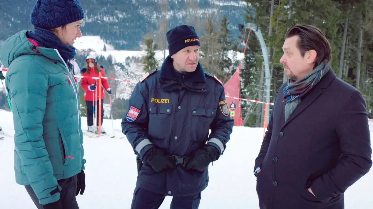 Polizist im Schnee, die Landschaft ist schwarz wie schnee. Eine Frau in t&uuml;rkiser Jacke und ein Mann im dunklen Mantel stehen daneben.