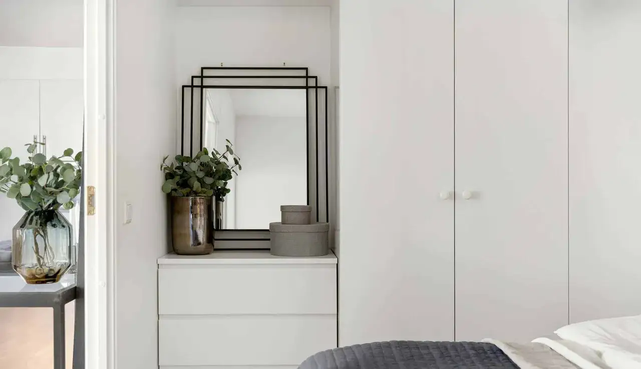 Ein moderner spiegel im schlafzimmer &uuml;ber einer wei&szlig;en Kommode mit Pflanzen und runden Boxen. Daneben ein gro&szlig;er wei&szlig;er Kleiderschrank.