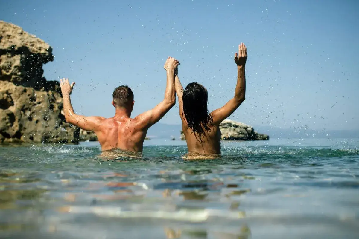 Pareja disfrutando de las playas nudistas Galicia, con los brazos en alto y salpicaduras de agua.