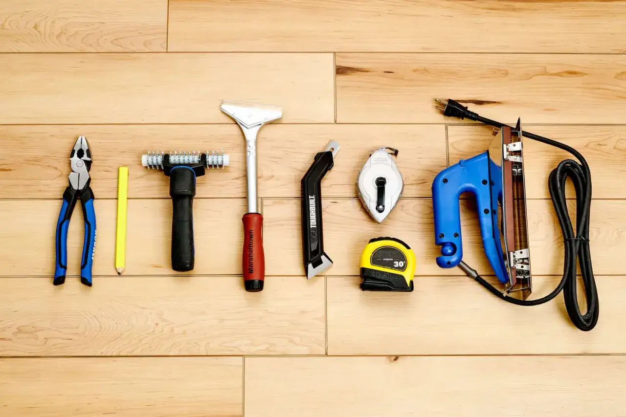 Werkzeuge für das **Teppich verlegen**: Zange, Bleistift, Teppichmesser, Maßband, Kreideleine und ein elektrischer Tacker liegen auf Holzdielen.