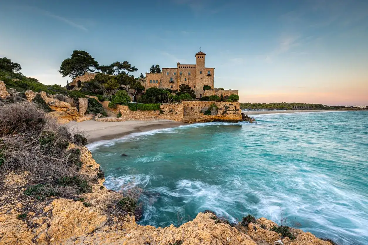 Ein Schloss thront &uuml;ber einem einsamen Strand an der K&uuml;ste Spaniens. T&uuml;rkisfarbenes Wasser umsp&uuml;lt die Felsen.