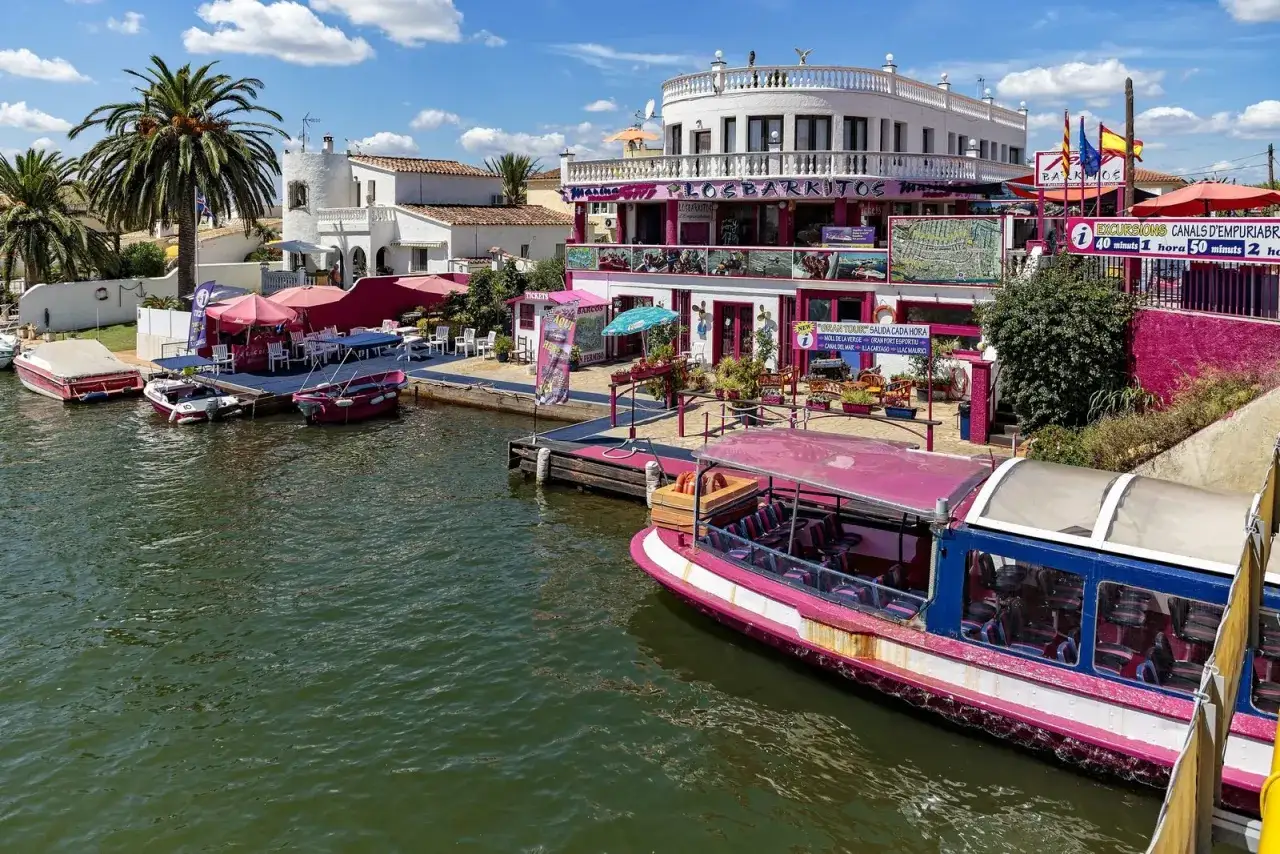 Bateaux roses et blancs amarrés le long des canaux d'Empuriabrava, Espagne. Un restaurant coloré avec des parasols roses et des palmiers complète le paysage.