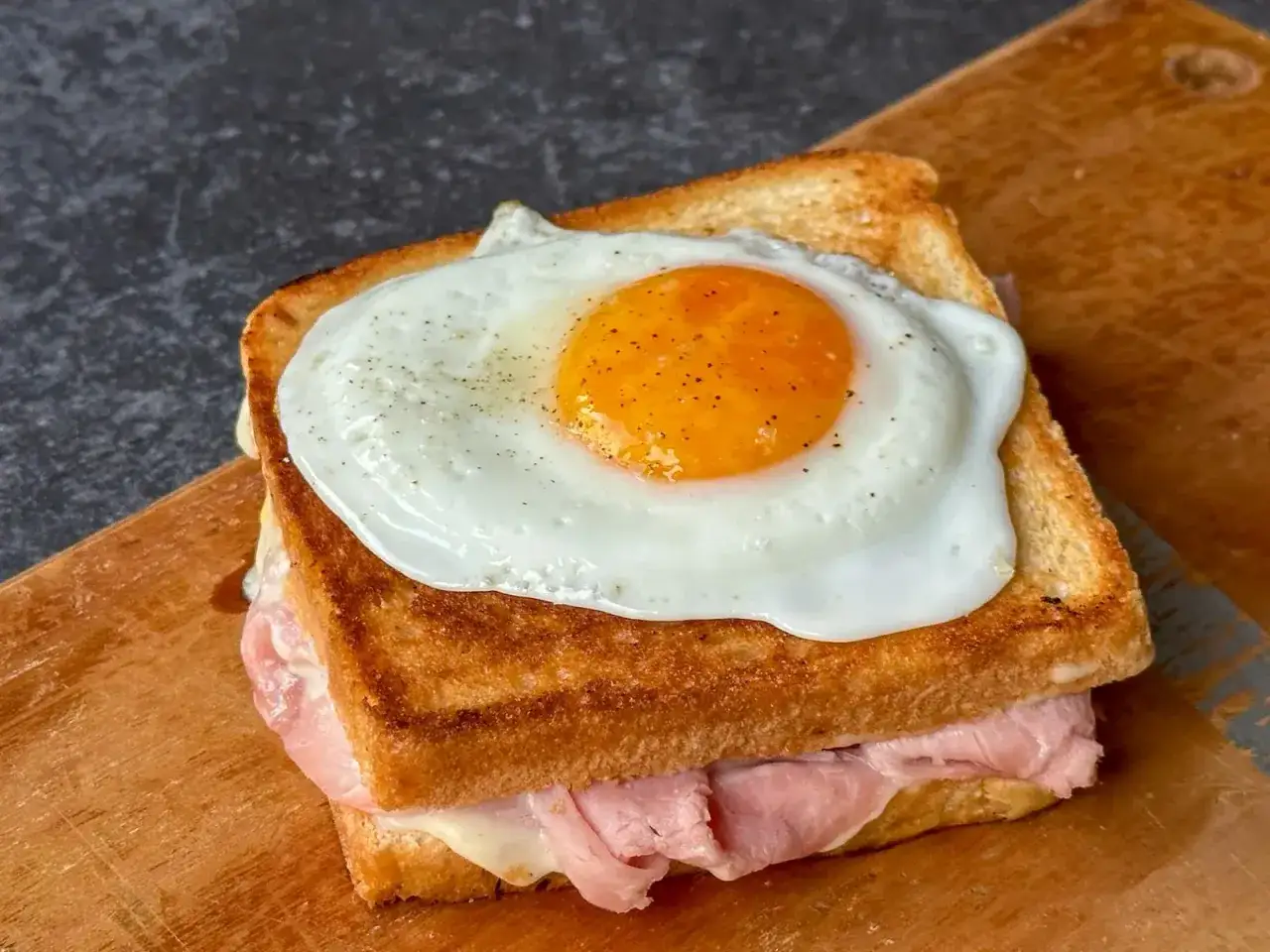 Ein k&ouml;stlicher Croque Madame mit Schinken, K&auml;se und einem perfekt gebratenen Spiegelei obendrauf, serviert auf einem Holzbrett.