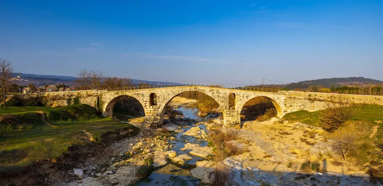 Le **pont Julien**, un ancien pont de pierre &agrave; trois arches, enjambe une rivi&egrave;re ass&eacute;ch&eacute;e bord&eacute;e de rochers et de v&eacute;g&eacute;tation.
