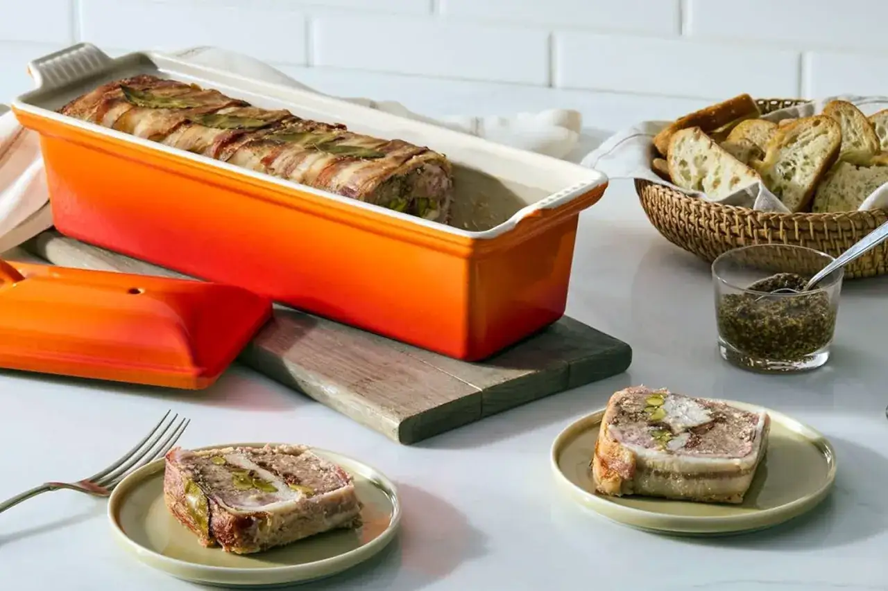 Terrina de manitas de cerdo envuelta en tocino, lista para hornear en un recipiente naranja. Servida en platos, acompa&ntilde;ada de pan y mostaza.