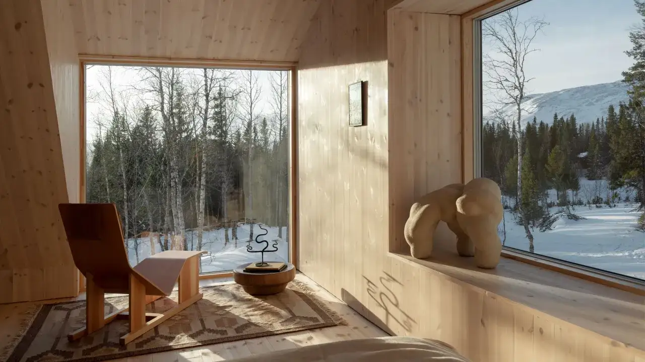 Int&eacute;rieur d'une maison dans les bois, avec de grandes fen&ecirc;tres offrant une vue sur une for&ecirc;t enneig&eacute;e et des montagnes. Un fauteuil design et une sculpture abstraite compl&egrave;tent le d&eacute;cor.