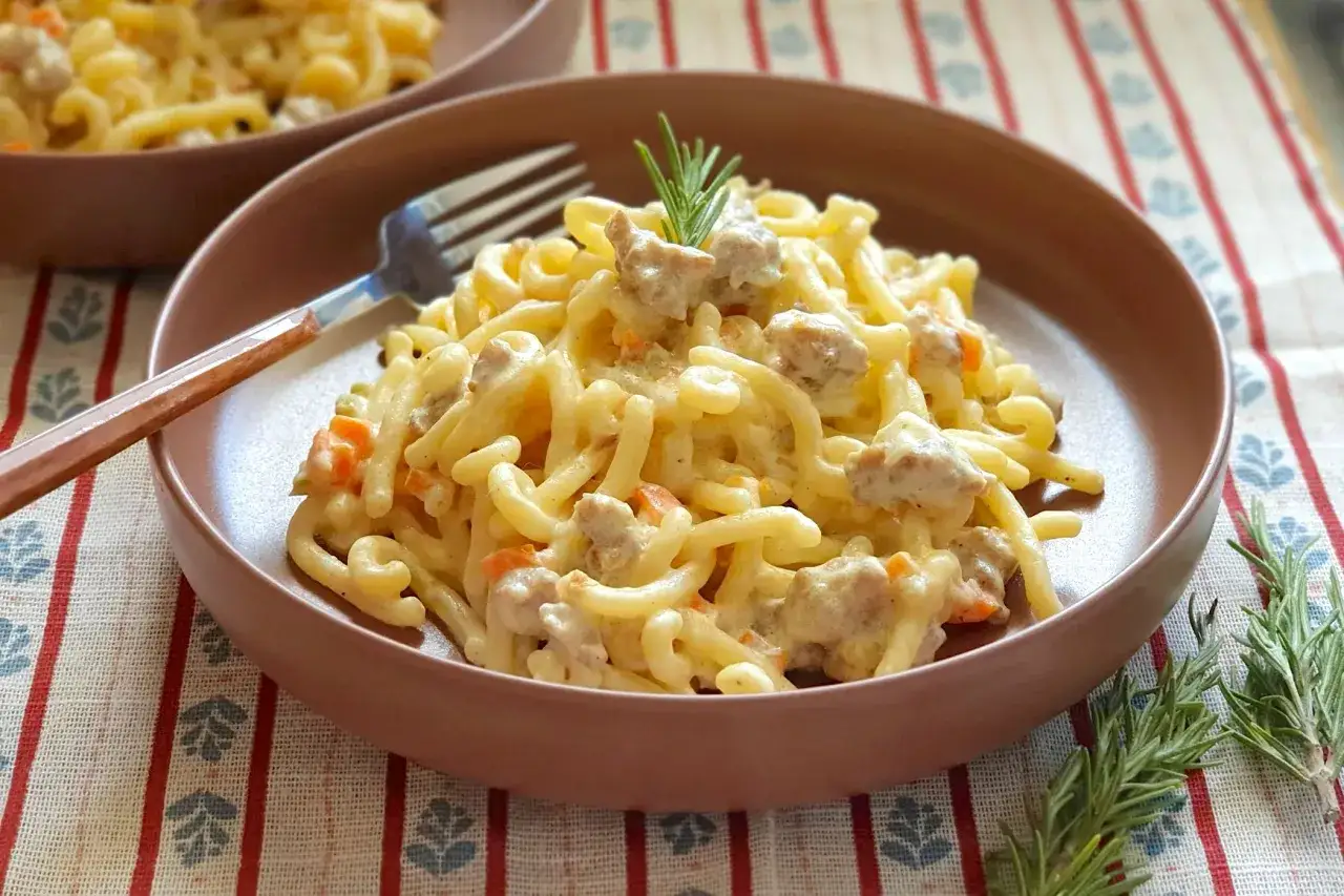 Un piatto di gramigna alla salsiccia, condita con una cremosa salsa e pezzetti di salsiccia e carote. Un rametto di rosmarino decora il tutto.