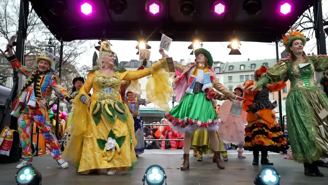 Bunte Kost&uuml;me und fr&ouml;hliche Gesichter auf einer B&uuml;hne, bereit f&uuml;r den Fasching 2026. Ein Mann im bunten Anzug, eine Frau im gelben Kleid mit Bananen, und weitere Darsteller in fantasievollen Outfits.