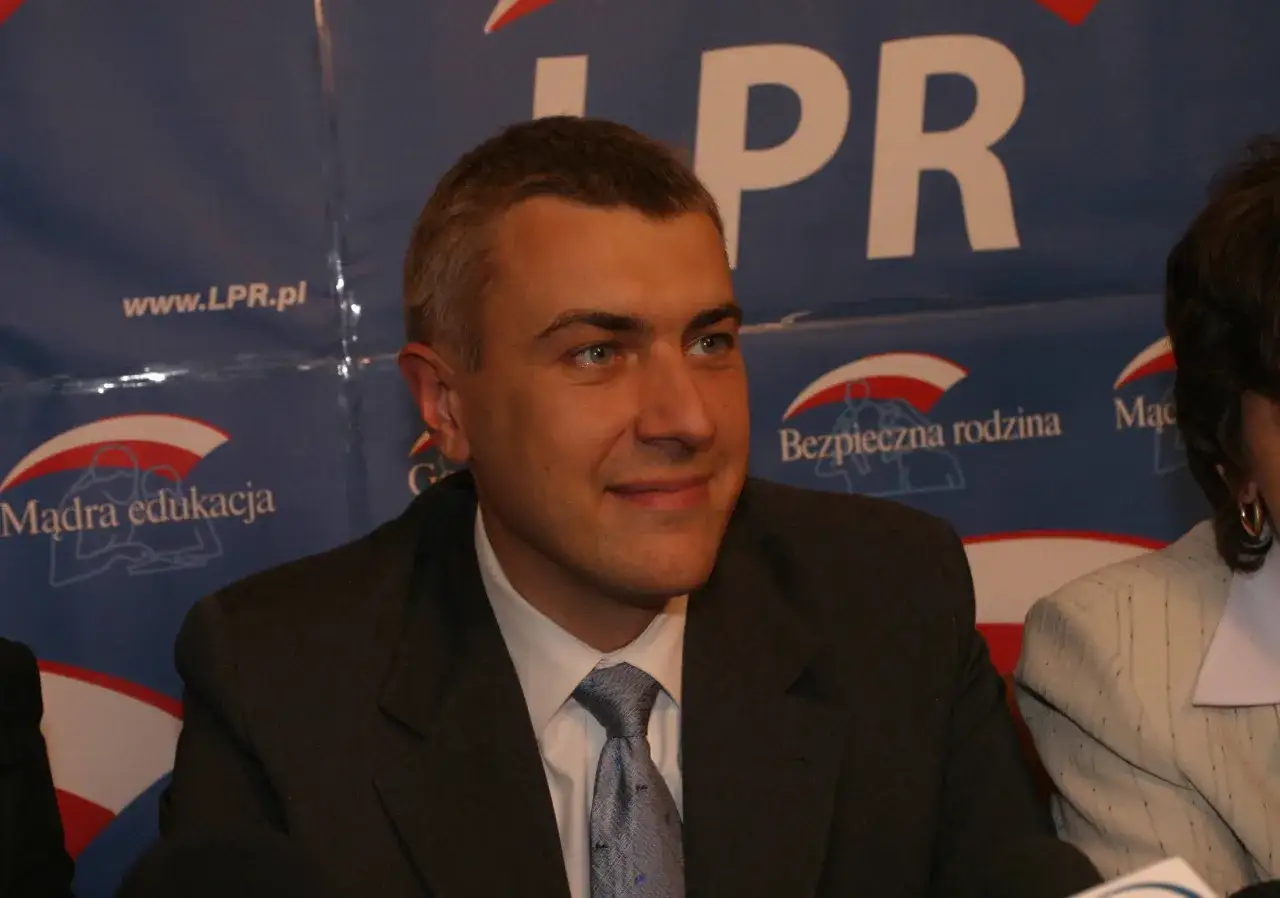 Mężczyzna w garniturze, z niebieskimi oczami, uśmiecha się na tle baneru LPR. W tle widać hasła 