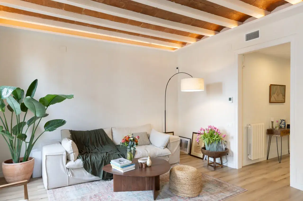 Salón acogedor tras reforma piso pequeño. Sofá beige, manta verde, mesa de centro de madera, plantas y flores. Vigas de madera en el techo.
