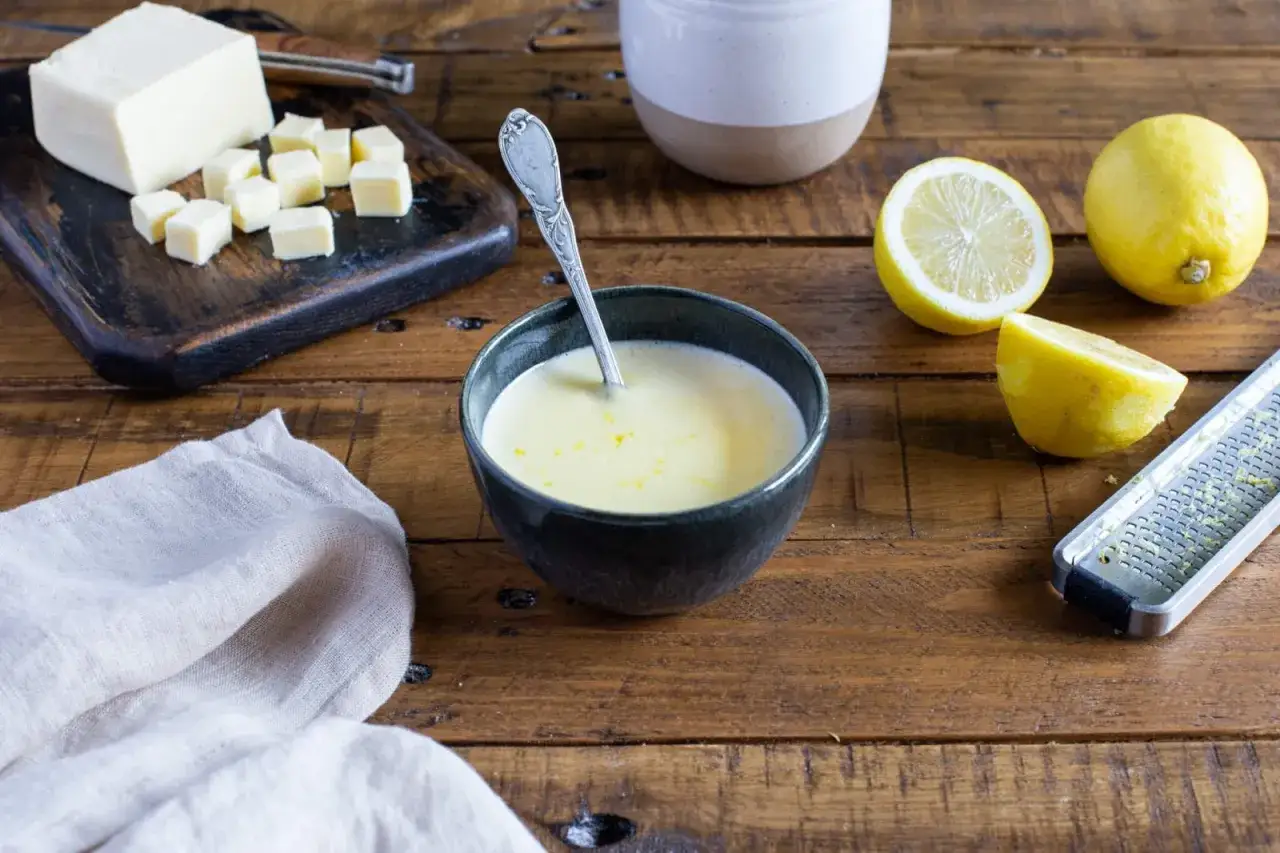 Un bol de sauce beurre citron, avec des citrons, du beurre coup&eacute; en d&eacute;s et une r&acirc;pe &agrave; zester sur une table en bois.