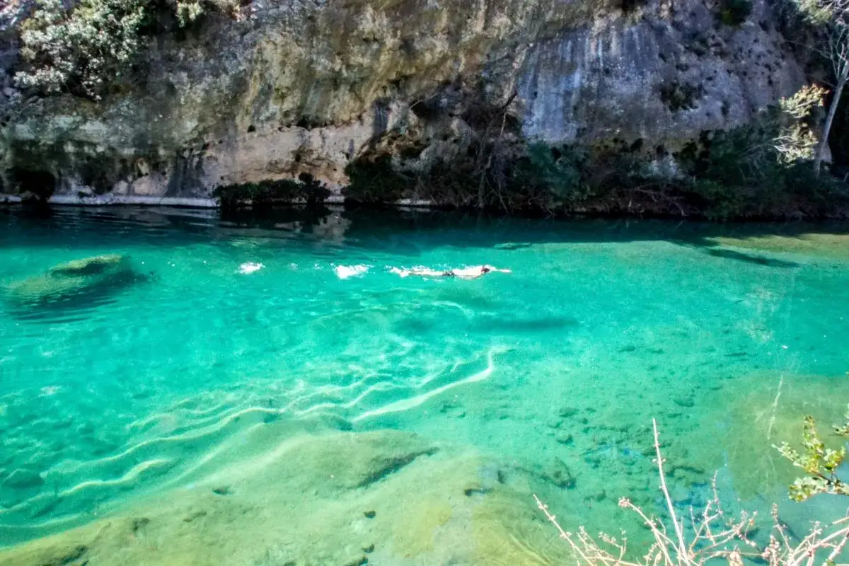 Persona nada en un pou clar de aguas turquesas, bajo un acantilado rocoso.