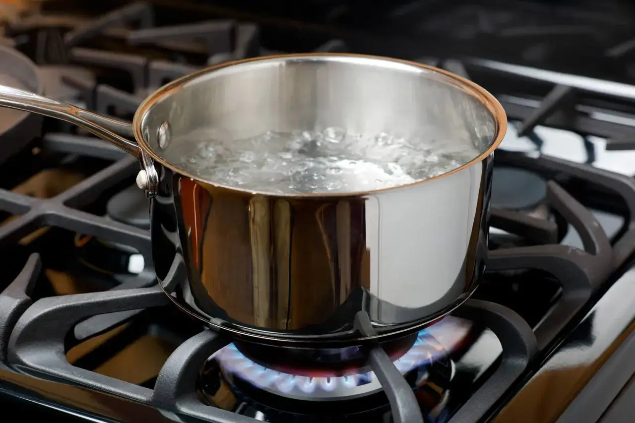 Une casserole en acier inoxydable sur une cuisinière à gaz, l'eau commence à **bouillir**. La flamme bleue illumine le fond de la casserole.