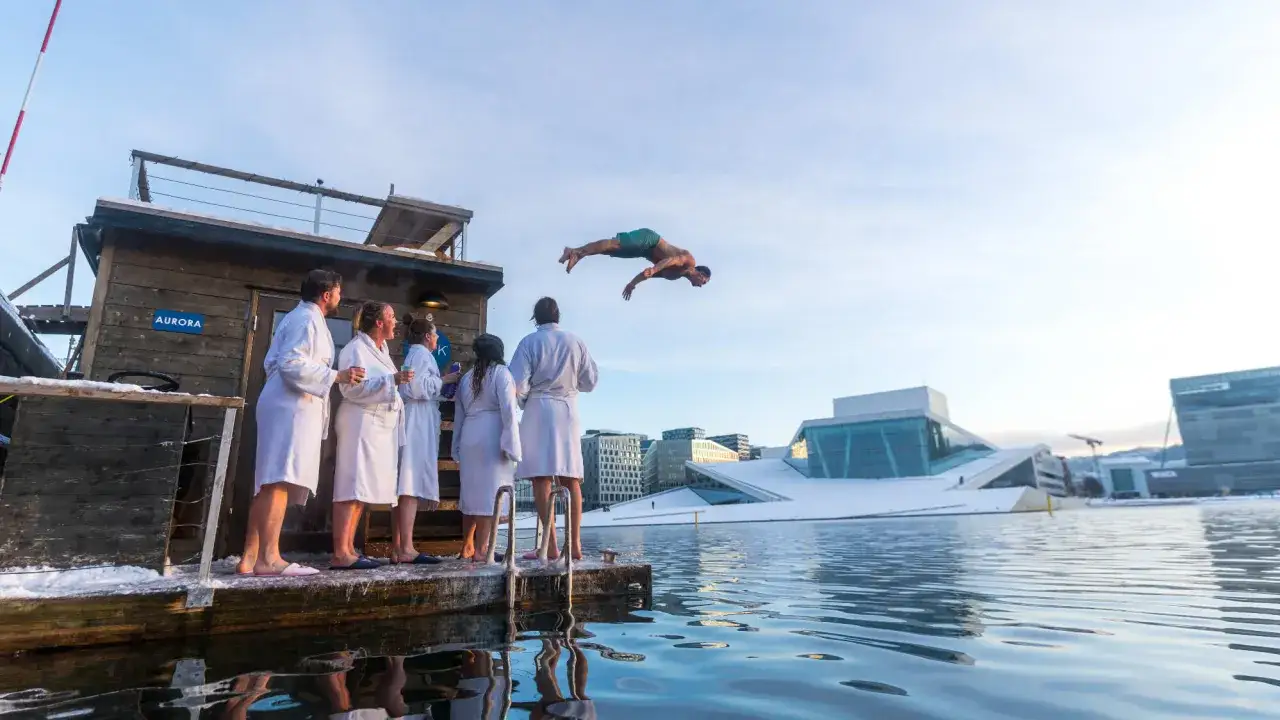 Ein Mann springt im **Oslo Winter** von einer Sauna ins eiskalte Wasser, w&auml;hrend Freunde in Badem&auml;nteln zusehen.
