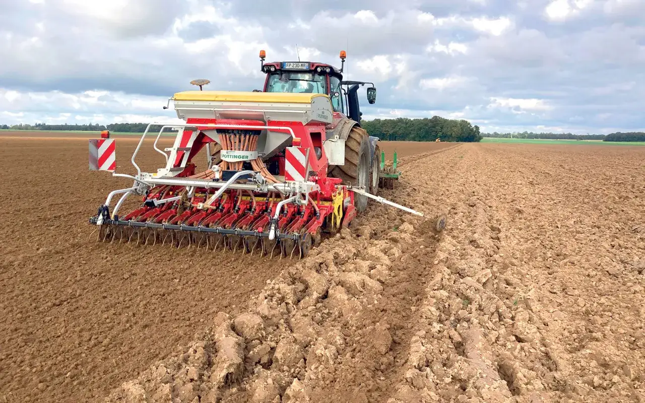 Un tracteur rouge sème le blé dans un champ labouré. Le semoir Pöttinger prépare la terre pour la future récolte.