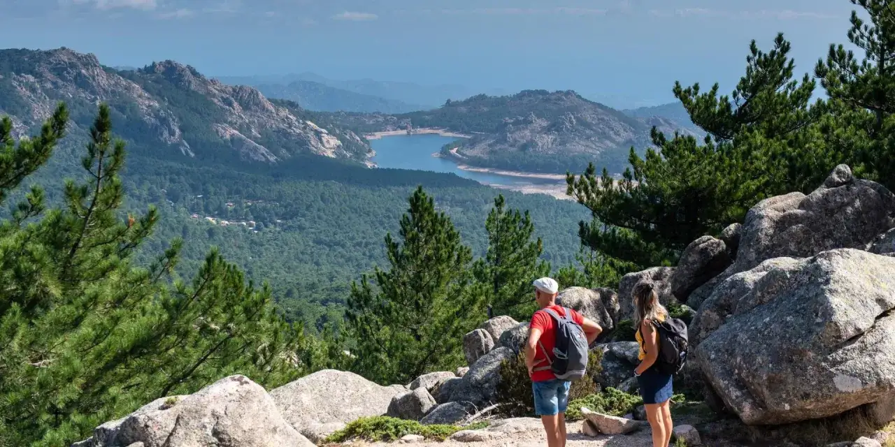 Deux randonneurs contemplent un lac en forme de croissant, la mare au centre du paysage, entouré de montagnes verdoyantes et de pins.