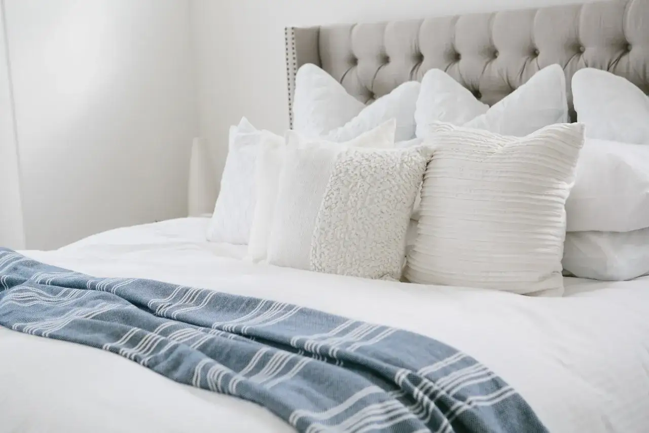 A bed with a tufted headboard, layered white pillows, and a blue striped throw. The big square pillows, called Euro shams, add a cozy touch.
