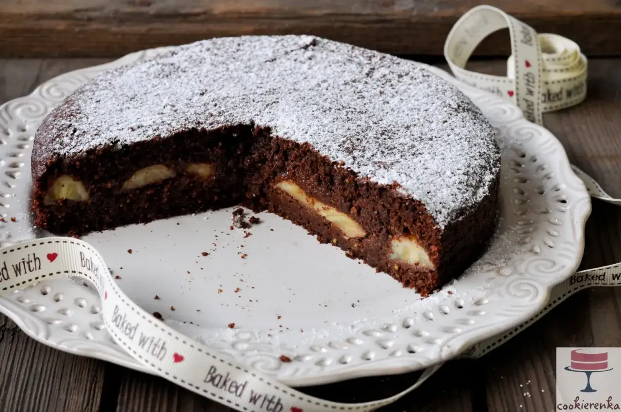 Czekoladowa babka jajowata z kawałkami jabłek, posypana cukrem pudrem, na talerzu z ozdobną wstążką.