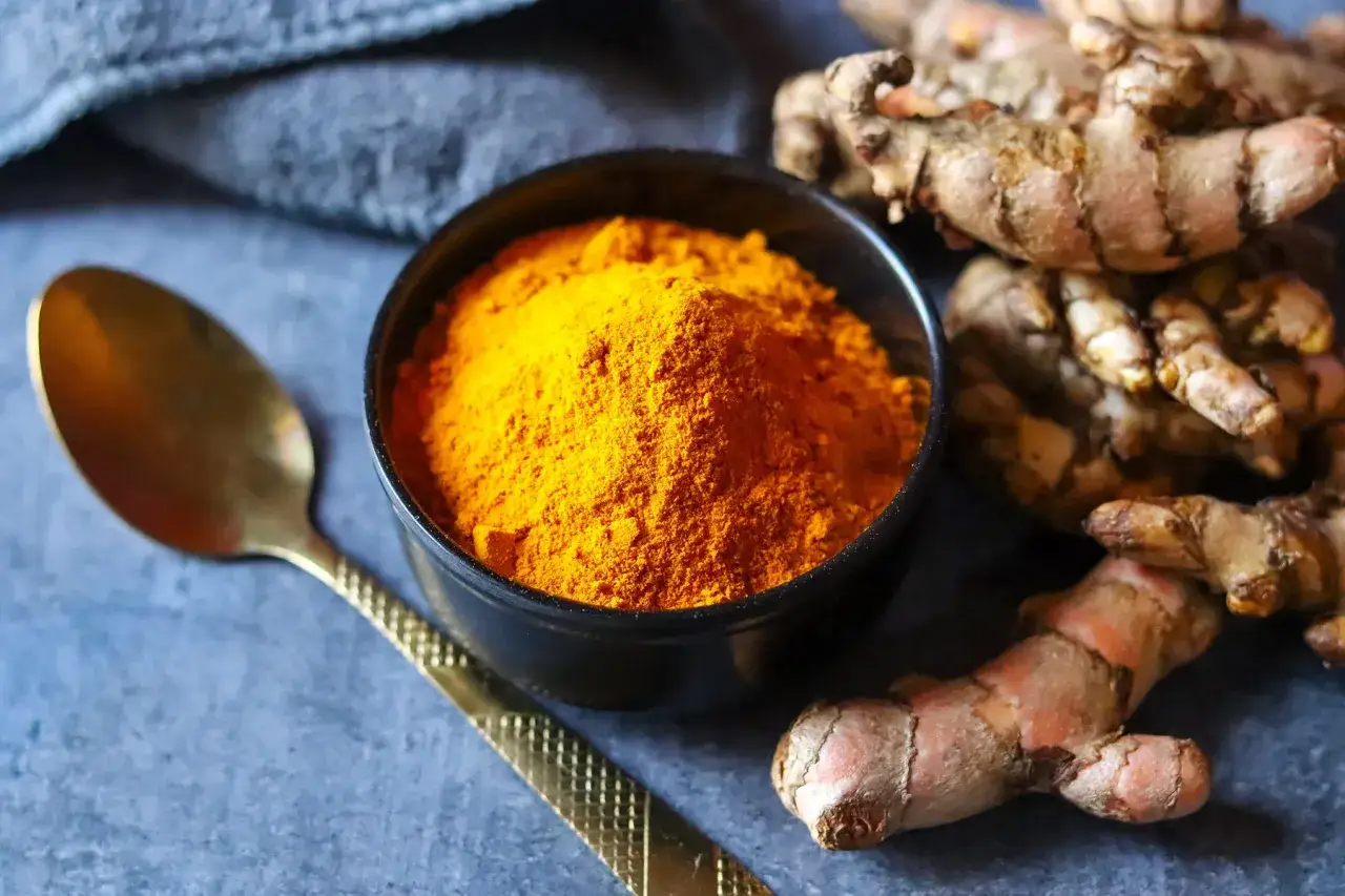 Poudre de curcuma dans un bol, à côté de racines de curcuma et d'une cuillère dorée. Attention au curcuma danger potentiel.