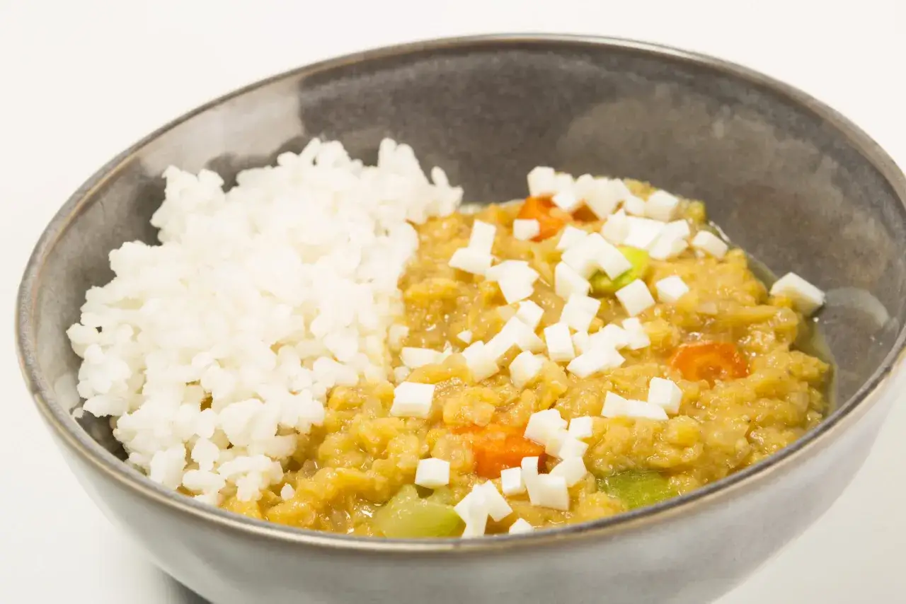 Un taz&oacute;n de lentejas rojas con arroz blanco, adornado con cubitos de queso y trozos de vegetales.