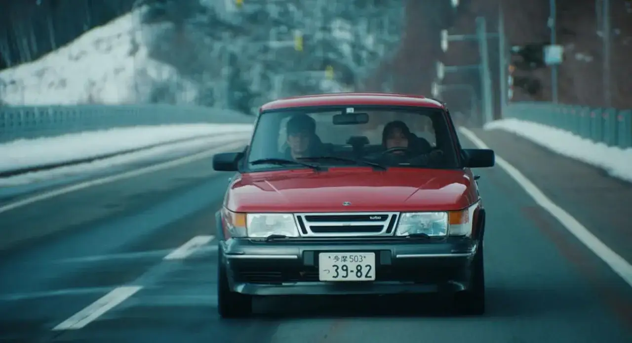 En röd Saab 900 Turbo körs på en snöig väg. En passagerare sitter bredvid föraren. En scen från en 