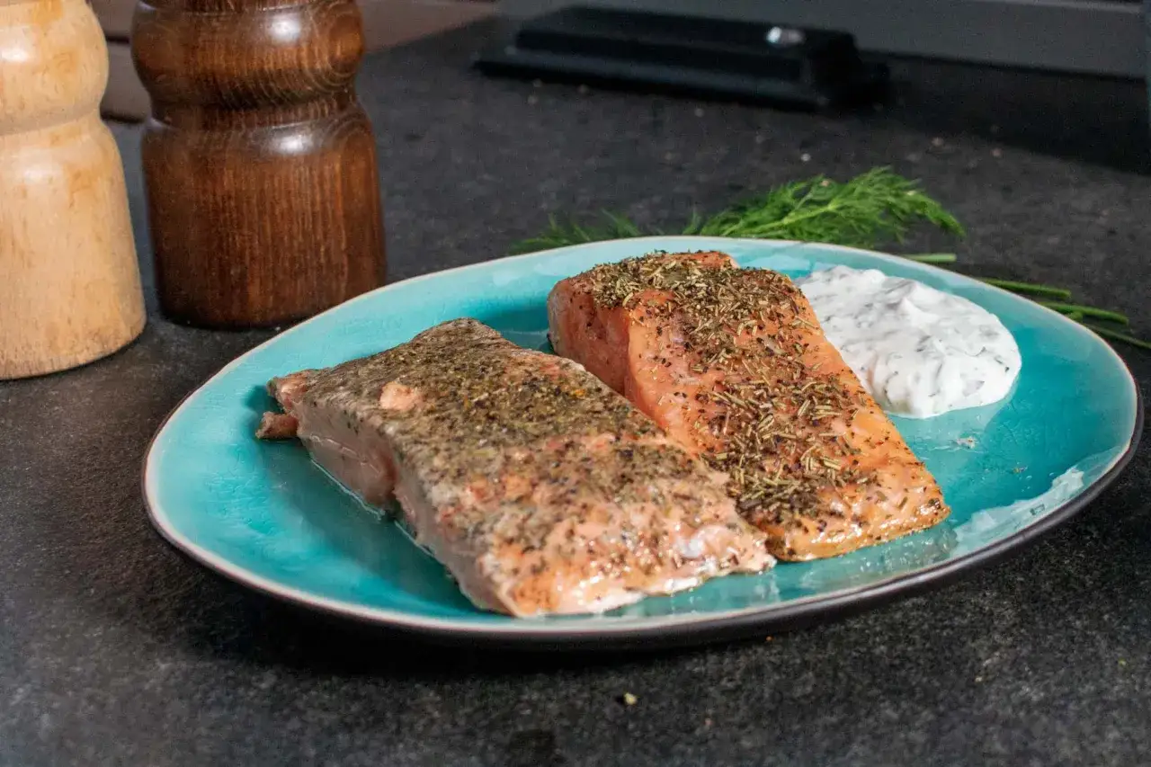 Zwei perfekt gegarte Lachs-Filets, zubereitet sous vide, liegen auf einem türkisfarbenen Teller, garniert mit Kräutern und einem Klecks Dip.