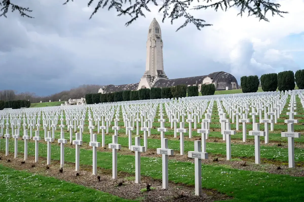 Reihen weißer Kreuze auf einem Friedhof, ein Denkmal im Hintergrund. Ein Ort des Gedenkens und des **dark tourism**.