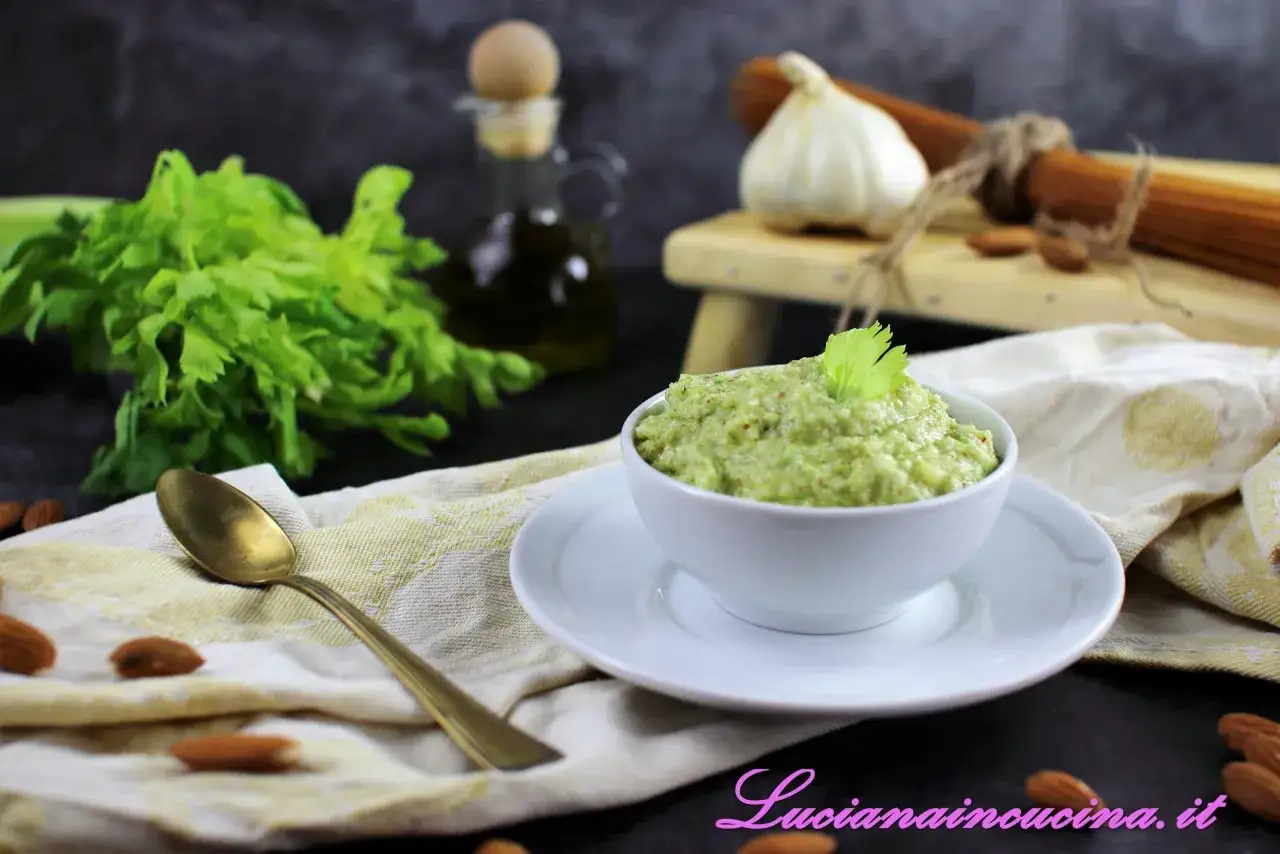 Un cremoso pesto di sedano, guarnito con una fogliolina verde, pronto per condire la pasta. Ingredienti freschi e mandorle completano la scena.