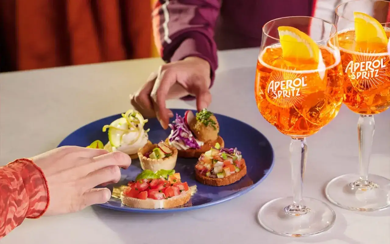 Zwei Aperol Spritz Gl&auml;ser und eine Auswahl an kleinen H&auml;ppchen auf einem blauen Teller. Perfekt f&uuml;r einen Aperitif!