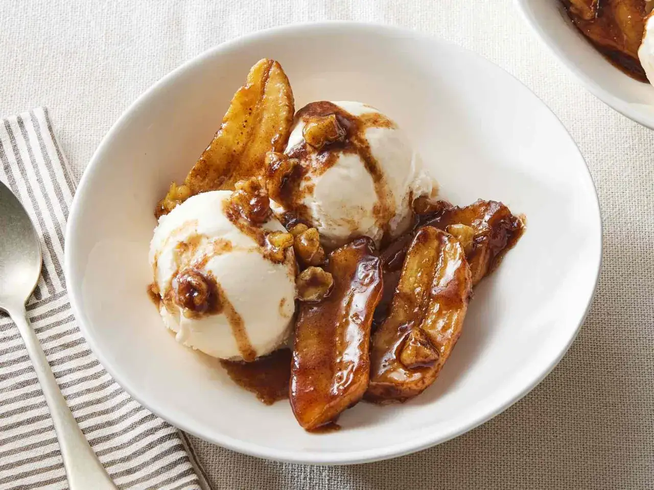 Zwei Kugeln Vanilleeis mit karamellisierter gebackener Banane und N&uuml;ssen in einer wei&szlig;en Sch&uuml;ssel.